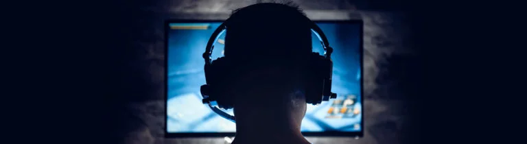 myty-o-programovani-banner-1920-525-768x210 Back view of a person wearing a gaming headset, facing a bright computer monitor in a dark room.