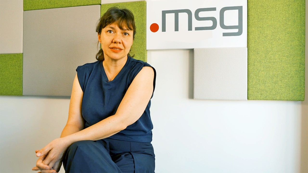Portrait of a woman in blue scrubs seated in front of a wall with green acoustic panels and a .msg logo behind her.