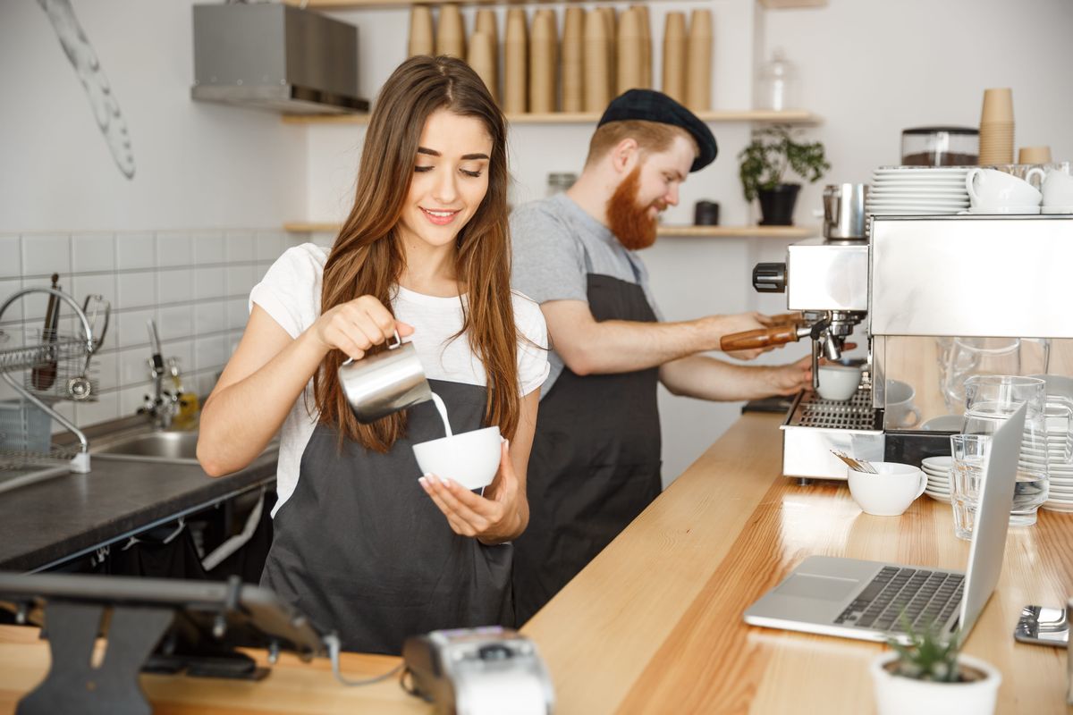 barista nalieva mlieko do šálky kávy v kuchyni a barista v pozadí pripravuje kávu v kávovare