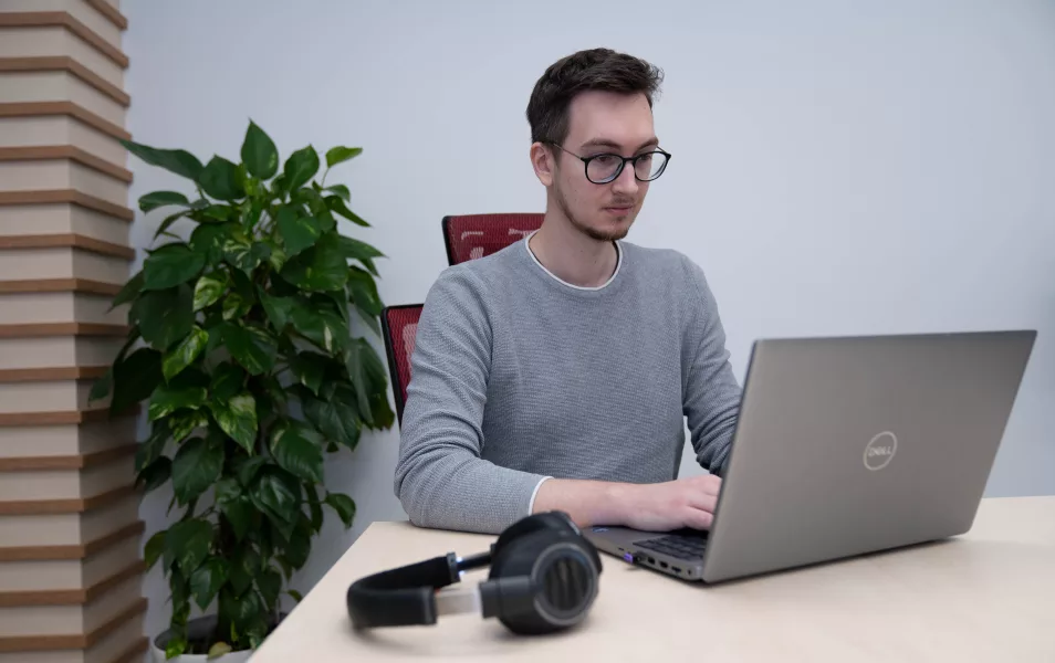 Java programátor Filip sedí v kancelárií na stoličke pri stole, kde je položený notebook a bezdrôtové slúchadlá.