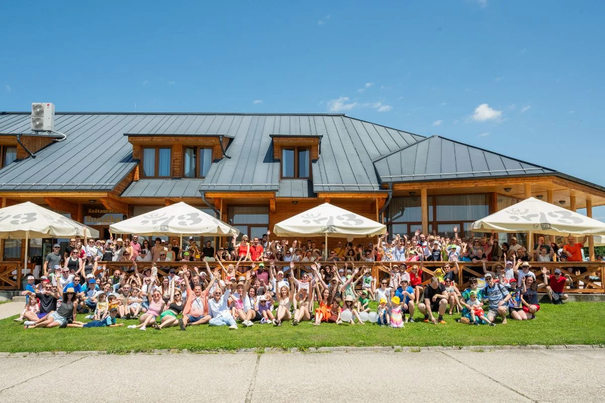 msg-firemny-teambuilding – msg life Slovakia Stovky ľudí s rodinami sedia a stoja pred drevenou chatou počas letného teambuildingu, mnohí mávajú do kamery.